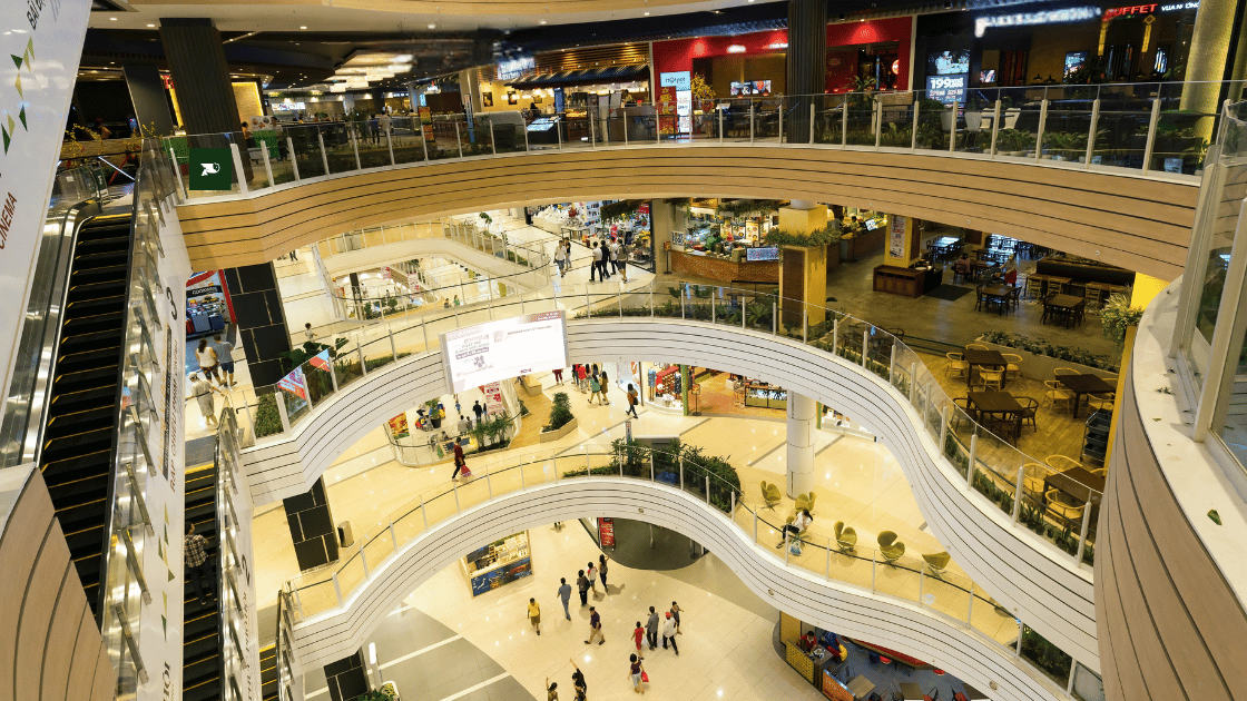 Interior view of a multi-level shopping mall with curved balconies, escalators, and various retail stores and restaurants. Shoppers are visible on all levels.