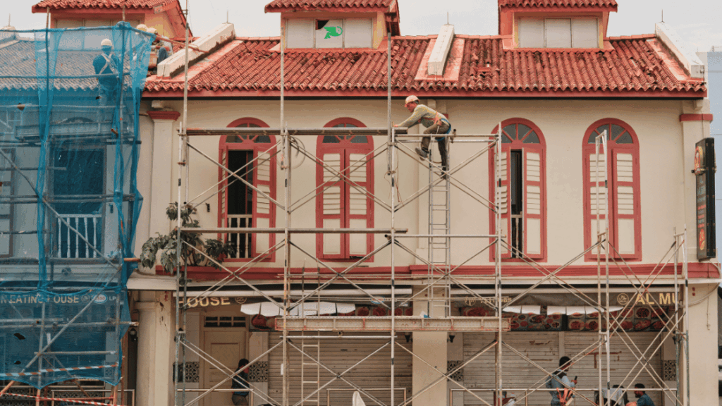 Worker climbing scaffolding in front of a red-roofed heritage building under renovation, with safety netting on one side and closed shopfronts below.
